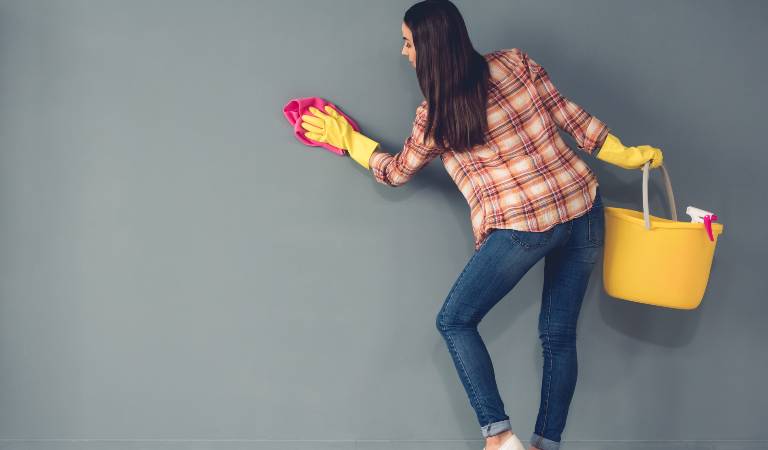 Woman in check shirt, blue jeans and yellow gloves cleaning wall with red cloth holding a yellow basket in her hand