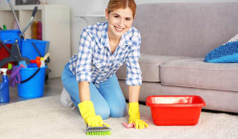 Woman in check shirt, blue jeans and yellow gloves cleaning carpet with a brush.