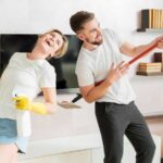 Woman holding a bottle and a man holding a stick dancing together in a clean home.