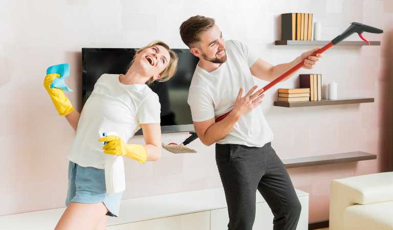 Woman holding a bottle and a man holding a stick dancing together in a clean home.