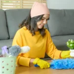 Young happy woman wearing yellow gloves and dusting the table in living room